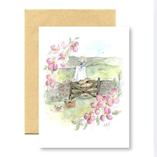 Greeting card with a watercolor illustration of a girl on a bridge surrounded by flowers, with a gold envelope.